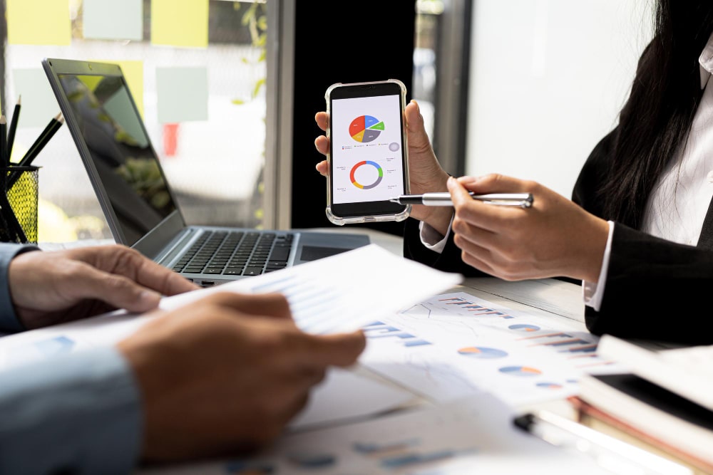 Deux personnes analysent des données financières sur un mobile et un ordinateur portable, illustrant les services d'une agence de marketing mobile