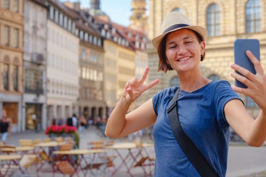 Une jeune femme souriante prend un selfie sur une place historique européenne, à la découverte de destinations insolites.