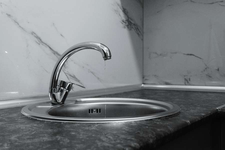 Un robinet goutte au-dessus d'un évier rond en inox installé dans un coin de cuisine avec un plan de travail sombre et un carrelage marbré blanc.